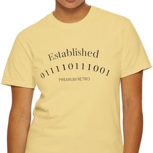 Może przedstawiać: Jasnożółta koszulka z napisem "Established" nad sekwencją kodu binarnego i "PREMIUM RETRO" poniżej. Koszulka ma klasyczny okrągły dekolt.