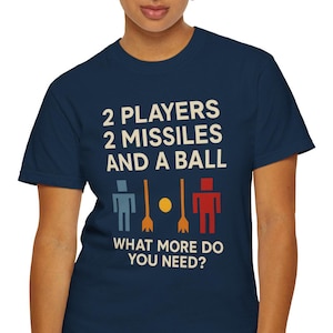Puede incluir: Camiseta azul marino con el texto "2 PLAYERS 2 MISSILES AND A BALL" en blanco. Debajo, tres figuras de palo y la pregunta "WHAT MORE DO YOU NEED?" en blanco. La camiseta está siendo usada.