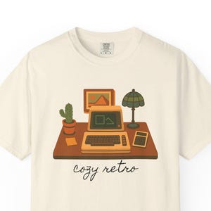 May include: Cream-colored t-shirt featuring a retro computer illustration with a cactus, lamp, and the words "cozy retro." The design includes a vintage computer, a small picture frame, and a calculator on a brown desk.