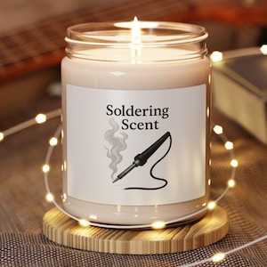 Peut inclure: Une bougie allumée dans un pot en verre transparent avec une étiquette blanche indiquant "Soldering Scent" et un fer à souder. La bougie est posée sur un petit dessous de verre rond en bois. Des guirlandes lumineuses et une guitare sont en arrière-plan.
