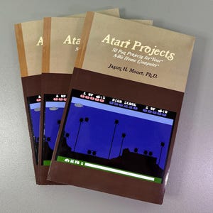Könnte beinhalten: Ein Stapel von vier braunen Büchern mit dem Titel "Atari Projects: 50 Fun Projects for Your 8-Bit Home Computer" von Jason H. Moore, Ph.D. Das Cover zeigt einen Screenshot eines Videospiels mit blauem Hintergrund, einem Raumschiff und Palmen.