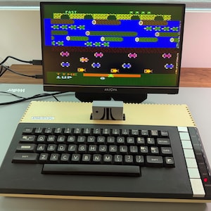 May include: An Atari 800XL computer with a connected monitor displaying a retro video game. The computer has a black keyboard and a beige case. The monitor is black and displays a colorful game scene.