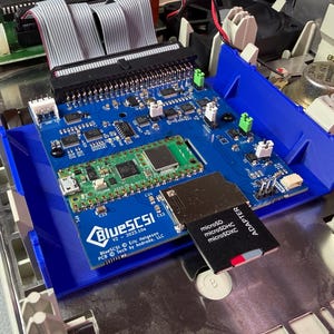 May include: A blue circuit board with various electronic components, including a green circuit board and a black adapter for a microSD card. The board is labeled "BlueSCSI" and is mounted inside a computer case.