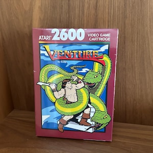 May include: Atari 2600 Venture video game cartridge box. The box features a muscular character fighting a large green snake. The game title "Venture" is displayed prominently with a blue background and red border.
