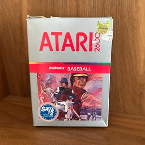 May include: An Atari 2600 video game cartridge box for RealSports Baseball. The box is silver with red text that reads "ATARI 2600" and "RealSports BASEBALL". The box art features baseball players in action.