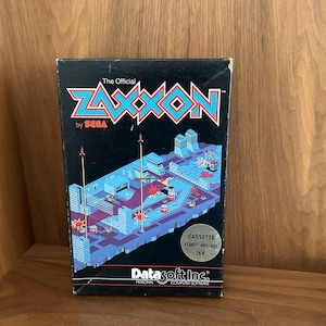 May include: A vintage video game cartridge box for "Zaxxon" by SEGA. The box is black with the game title in teal and red. The box art depicts a top-down view of a futuristic cityscape with a pixelated aesthetic. The box also includes the DataSoft Inc. logo.