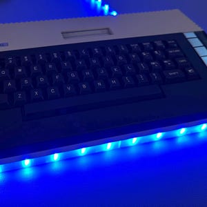 May include: An Atari 800XL computer with a black keyboard and a beige top. The computer is illuminated with blue LED lights along the bottom edge. The Atari logo is visible on the top.