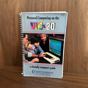 May include: A spiral-bound book titled "Personal Computing on the VIC-20" with a colorful logo. The cover features a family gathered around a computer. The book is a "friendly computer guide" from Commodore Computer.
