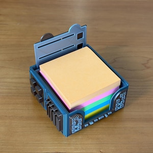 May include: A blue and brown decorative desk organizer with a stack of colorful sticky notes inside. The organizer has a hinged top and a geometric design on the sides. The sticky notes are in shades of yellow, pink, green, and blue.