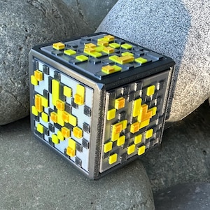May include: A pixelated cube with a silver frame, black top and bottom, and yellow and gray accents. The design resembles a block from a video game. The cube is sitting on a bed of gray rocks.