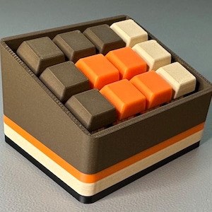 May include: A brown, rectangular container with a layered design of black, white, and orange stripes. The container holds several square-shaped blocks in shades of brown, orange, and off-white. The blocks are arranged in a grid pattern.