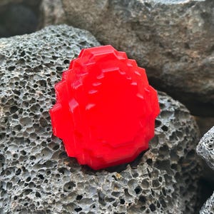 May include: A bright red, pixelated, gem-shaped object rests on a textured gray rock surface. The gem has a faceted design, catching the light and creating highlights. The overall aesthetic is reminiscent of retro video game graphics.