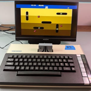 May include: An Atari 800 XE computer with a connected monitor displaying a retro video game. The keyboard is black with white lettering. The monitor is black and has the brand name "ARZOPA" on the bottom.