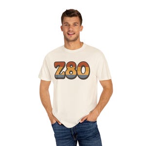 May include: Cream-colored t-shirt with the word "Z80" in a retro font. The text is in shades of brown, orange, and blue, with a dark outline. The shirt is a casual style.