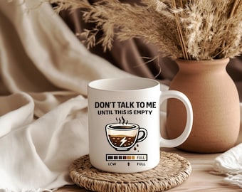May include: White ceramic coffee mug with the text "DON'T TALK TO ME UNTIL THIS IS EMPTY" and a coffee cup graphic. The mug sits on a round, woven coaster. A vase with dried plants is in the background.