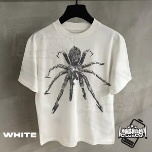 May include: White t-shirt featuring a large, detailed black spider graphic. The shirt has short sleeves and a crew neck. The word "WHITE" is printed at the bottom left. The "LowBatteryClubCo" logo is on the bottom right.