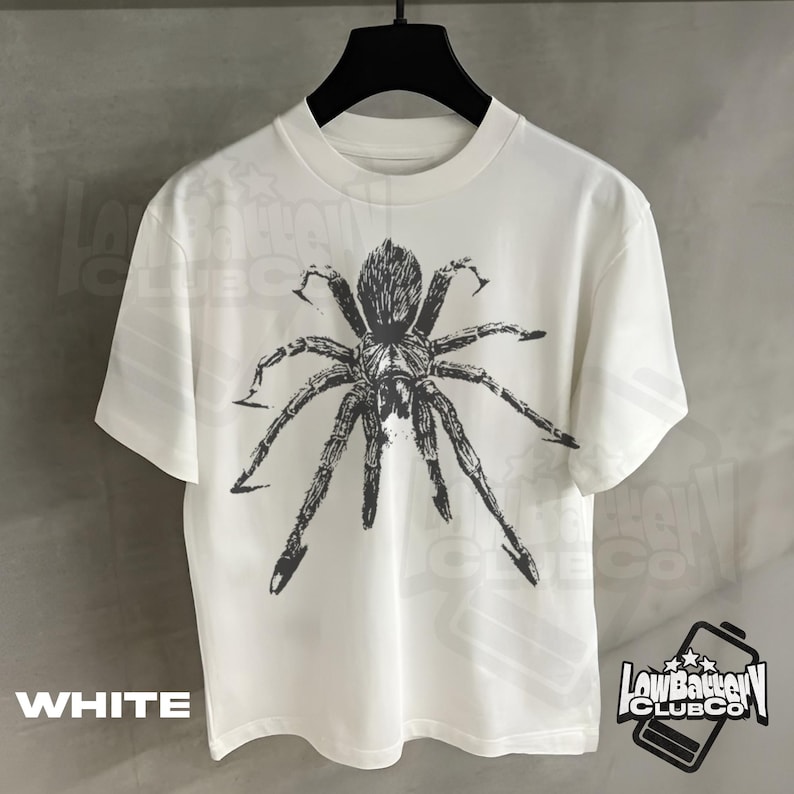 May include: White t-shirt featuring a large, detailed black spider graphic. The shirt has short sleeves and a crew neck. The word "WHITE" is printed at the bottom left. The "LowBatteryClubCo" logo is on the bottom right.