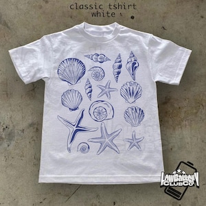 May include: White classic t-shirt with a blue seashell and starfish design. The shirt features various seashell illustrations and starfish. The text "classic tshirt white" is at the top.