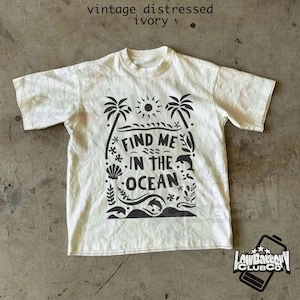 Puede incluir: Camiseta de color marfil con un dise&ntilde;o gr&aacute;fico negro que presenta palmeras, un sol, olas y el texto "FIND ME IN THE OCEAN". La camiseta tiene un aspecto vintage y desgastado, y cuello redondo.