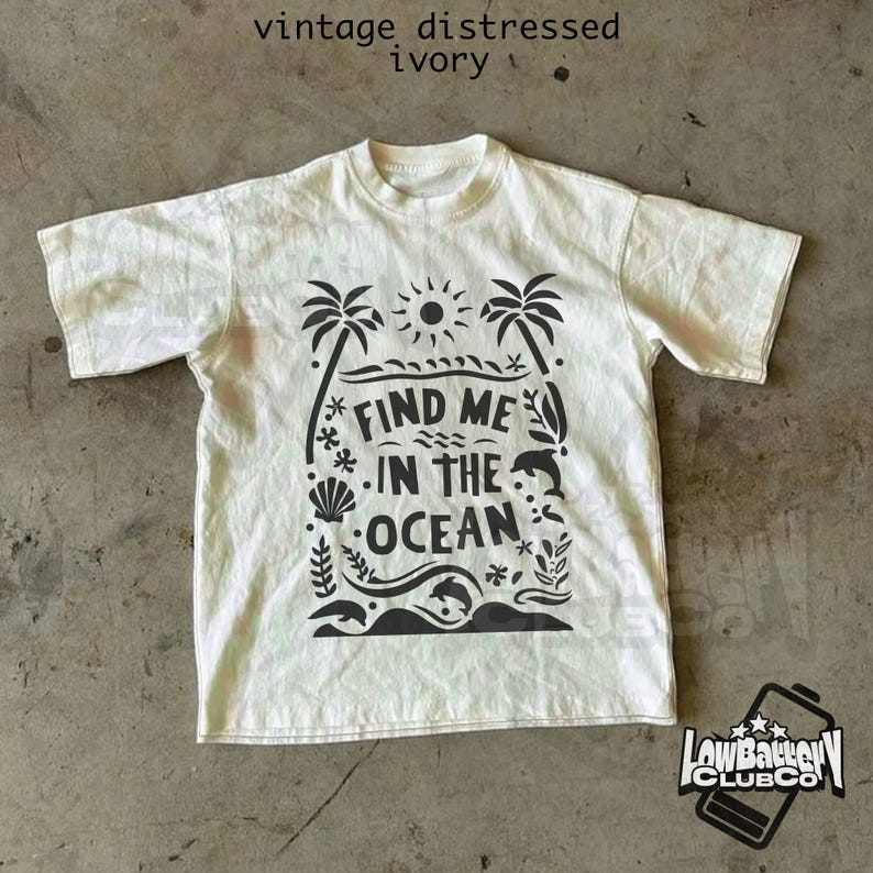 Puede incluir: Camiseta de color marfil con un dise&ntilde;o gr&aacute;fico negro que presenta palmeras, un sol, olas y el texto "FIND ME IN THE OCEAN". La camiseta tiene un aspecto vintage y desgastado, y cuello redondo.