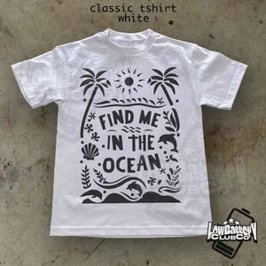 Puede incluir: Camiseta blanca cl&aacute;sica con un dise&ntilde;o gr&aacute;fico negro. El dise&ntilde;o incluye palmeras, un sol, olas, delfines y el texto "FIND ME IN THE OCEAN". La camiseta tambi&eacute;n tiene el texto "classic tshirt white" en la parte superior.