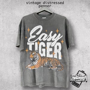 May include: A gray t-shirt with a distressed vintage look featuring a tiger and the text "Easy Tiger" in white with gold stars.