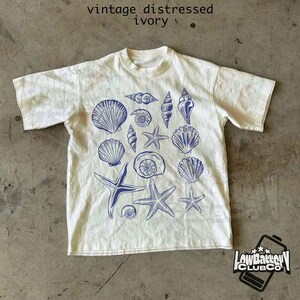 May include: Ivory-colored t-shirt with a crew neck and short sleeves. The shirt features a blue illustration of various seashells and starfish. The text "vintage distressed ivory" is at the top. The logo for "Low Battery Club Co" is at the bottom.