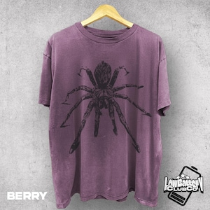 May include: Berry-colored t-shirt with a large black spider graphic. The shirt is hanging on a wooden hanger against a gray wall. The text "BERRY" is at the bottom left, and a logo is at the bottom right.