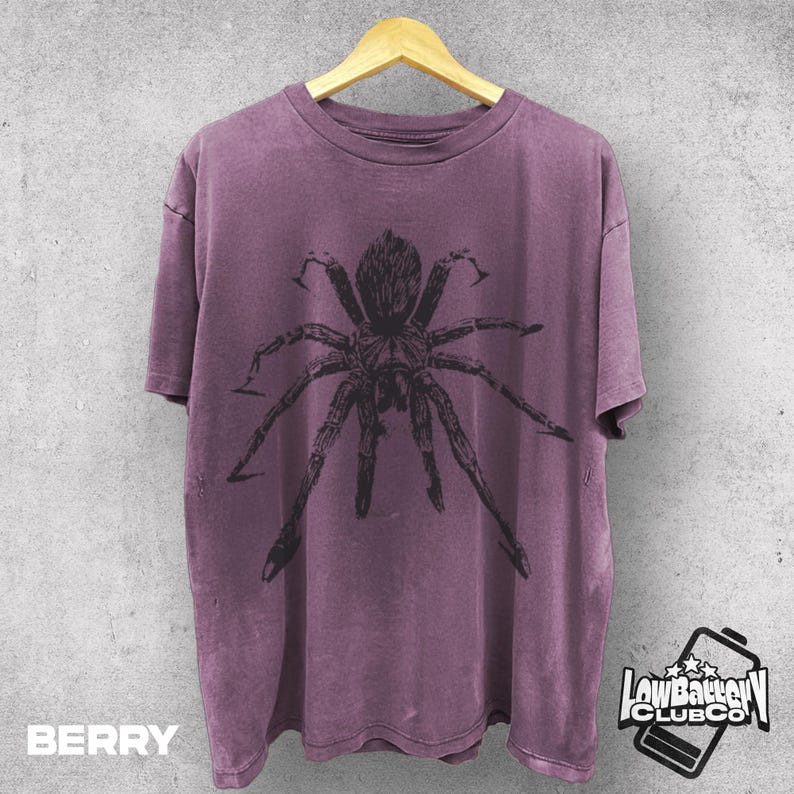 May include: Berry-colored t-shirt with a large black spider graphic. The shirt is hanging on a wooden hanger against a gray wall. The text "BERRY" is at the bottom left, and a logo is at the bottom right.