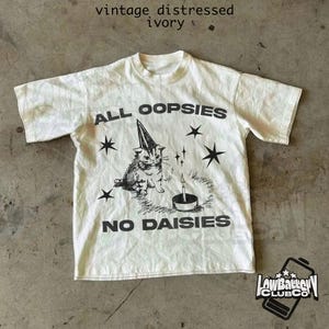 May include: A white vintage distressed t-shirt with a black graphic print of a cat wearing a party hat and the text "All Oopsies No Daisies".