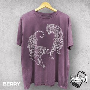 Peut inclure: T-shirt de couleur baie avec un motif de tigre blanc. Le t-shirt présente deux tigres face à face, avec le texte "BERRY" en bas à gauche. Le t-shirt est suspendu à un cintre en bois.