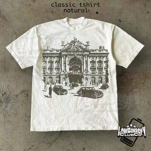 Puede incluir: Una camiseta clásica de color natural con un gráfico en blanco y negro de un edificio y coches antiguos. El texto "classic tshirt natural" está en la parte superior. La camiseta tiene cuello redondo y mangas cortas. El logotipo de la marca está en la parte inferior derecha.
