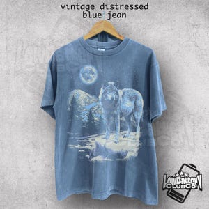 May include: A vintage blue jean t-shirt with a distressed look. The shirt features a graphic print of a wolf howling at the moon in a snowy forest scene.