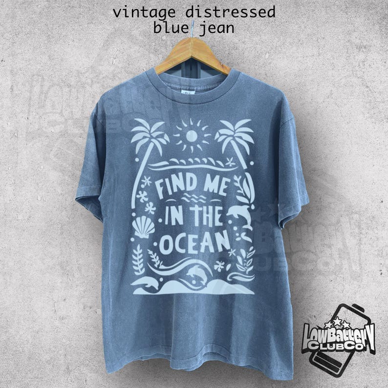 Puede incluir: Camiseta azul vaquero con un gr&aacute;fico blanco que dice "FIND ME IN THE OCEAN". El dise&ntilde;o incluye palmeras, un sol, olas y vida marina. La camiseta est&aacute; colgada en una percha de madera.
