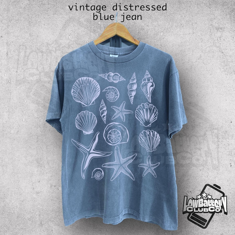 May include: Blue jean-colored t-shirt with a white seashell and starfish design. The shirt is hanging on a wooden hanger. The text "vintage distressed blue jean" is at the top.