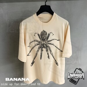 May include: Banana-colored t-shirt featuring a large, detailed spider graphic in dark gray. The shirt has short sleeves and a crew neck. The text "BANANA" and "size up for oversized fit" are printed at the bottom. The "LowBattery Club Co" logo is on the right.
