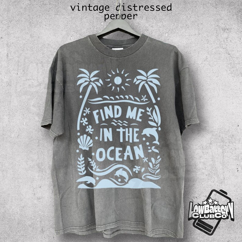 Puede incluir: Camiseta gris con un gr&aacute;fico azul claro que dice "Find Me In The Ocean". El dise&ntilde;o incluye palmeras, un sol, olas, una concha y un delf&iacute;n. La camiseta tiene un aspecto vintage y desgastado.