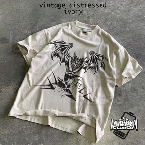 May include: Ivory-colored t-shirt with a distressed vintage look. The shirt features a black graphic of a bat-like creature with outstretched wings. The text "vintage distressed ivory" is printed at the top. The Low Battery Club Co. logo is at the bottom.