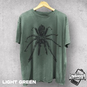 May include: Light green t-shirt with a large black spider graphic. The shirt is hanging on a wooden hanger. The text "LIGHT GREEN" is at the bottom of the shirt. The "Low Battery Club Co" logo is on the bottom right.