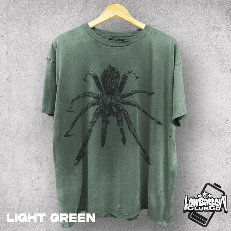 May include: Light green t-shirt with a large black spider graphic. The shirt is hanging on a wooden hanger. The text "LIGHT GREEN" is at the bottom of the shirt. The "Low Battery Club Co" logo is on the bottom right.