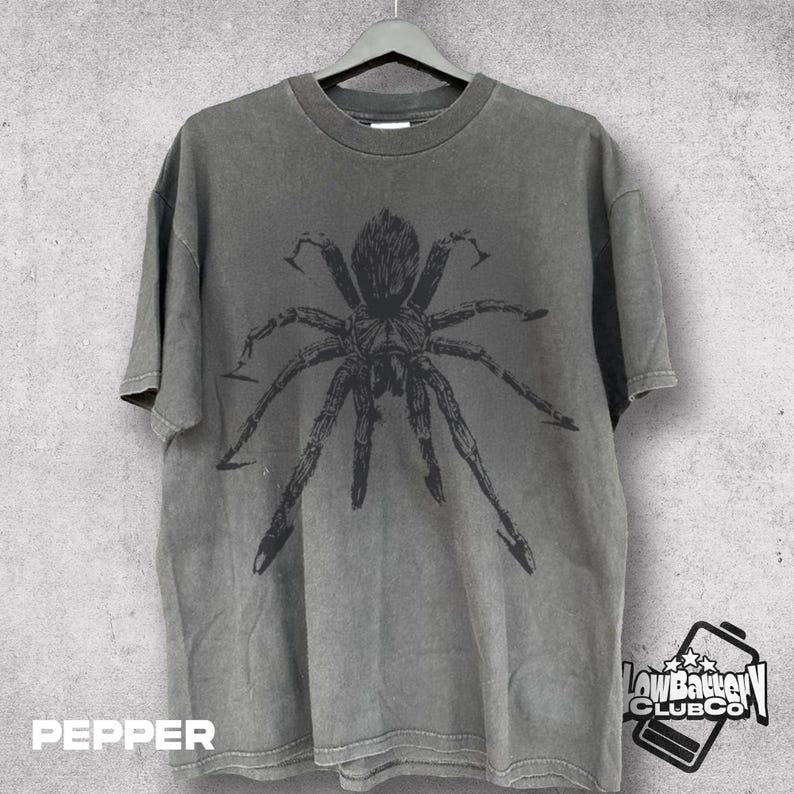 May include: Pepper-colored t-shirt with a large, detailed black spider graphic. The shirt is hanging on a black hanger against a gray background. The text "PEPPER" is at the bottom left.