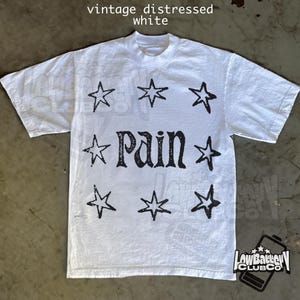 May include: White t-shirt with a crew neck featuring a distressed black graphic of the word "PAIN" surrounded by eight stars. The shirt has the text "vintage distressed white" at the top and a logo at the bottom.