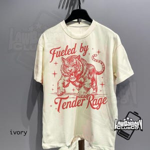 May include: Ivory-colored t-shirt featuring a red illustration of a tiger with the text "Fueled by Tender Rage." The shirt is hanging on a black hanger. The brand logo is visible on the bottom right.