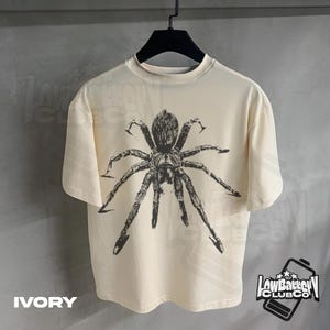 May include: Ivory-colored t-shirt featuring a large, detailed black spider graphic. The shirt has a crew neck and short sleeves. The text "IVORY" is at the bottom left. The "Low Battery Club Co" logo is at the bottom right.