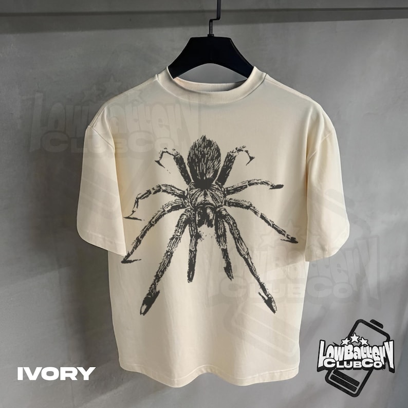 May include: Ivory-colored t-shirt featuring a large, detailed black spider graphic. The shirt has a crew neck and short sleeves. The text "IVORY" is at the bottom left. The "Low Battery Club Co" logo is at the bottom right.