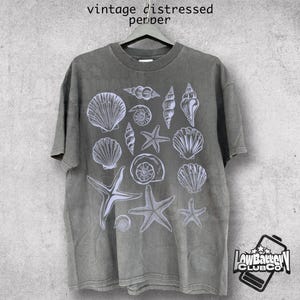 May include: A gray, vintage-style t-shirt featuring a white illustration of various seashells and starfish. The shirt has a distressed look, with the words "vintage distressed pepper" printed at the top. The shirt is hanging on a hanger.