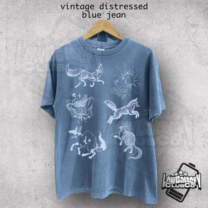May include: Blue jean-colored t-shirt with a distressed vintage look. The shirt features white illustrations of foxes and suns. The text "vintage distressed blue jean" is printed at the top. The shirt is hanging on a wooden hanger.