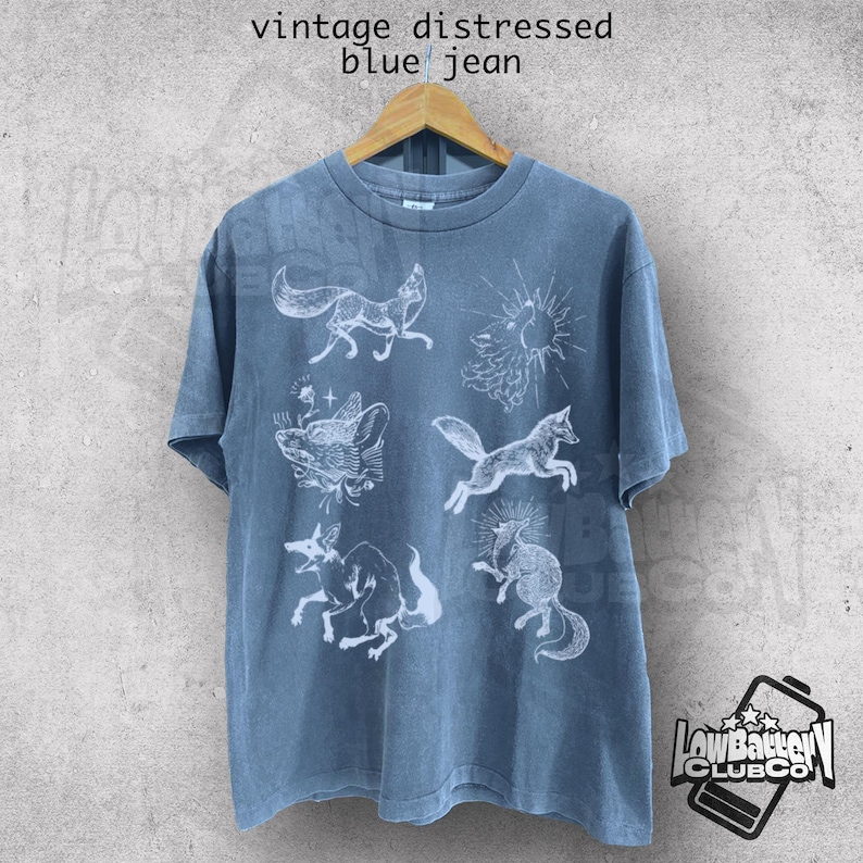 May include: Blue jean-colored t-shirt with a distressed vintage look. The shirt features white illustrations of foxes and suns. The text "vintage distressed blue jean" is printed at the top. The shirt is hanging on a wooden hanger.