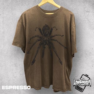 May include: Espresso-colored t-shirt featuring a large, detailed spider graphic. The shirt is hanging on a wooden hanger against a textured gray wall. The text "ESPRESSO" is at the bottom left.