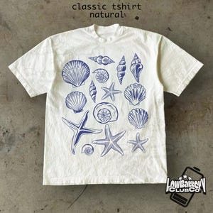 May include: A white t-shirt with a crew neck featuring a blue illustration of various seashells and starfish. The text "classic tshirt natural" is printed at the top. The shirt has short sleeves and a relaxed fit.